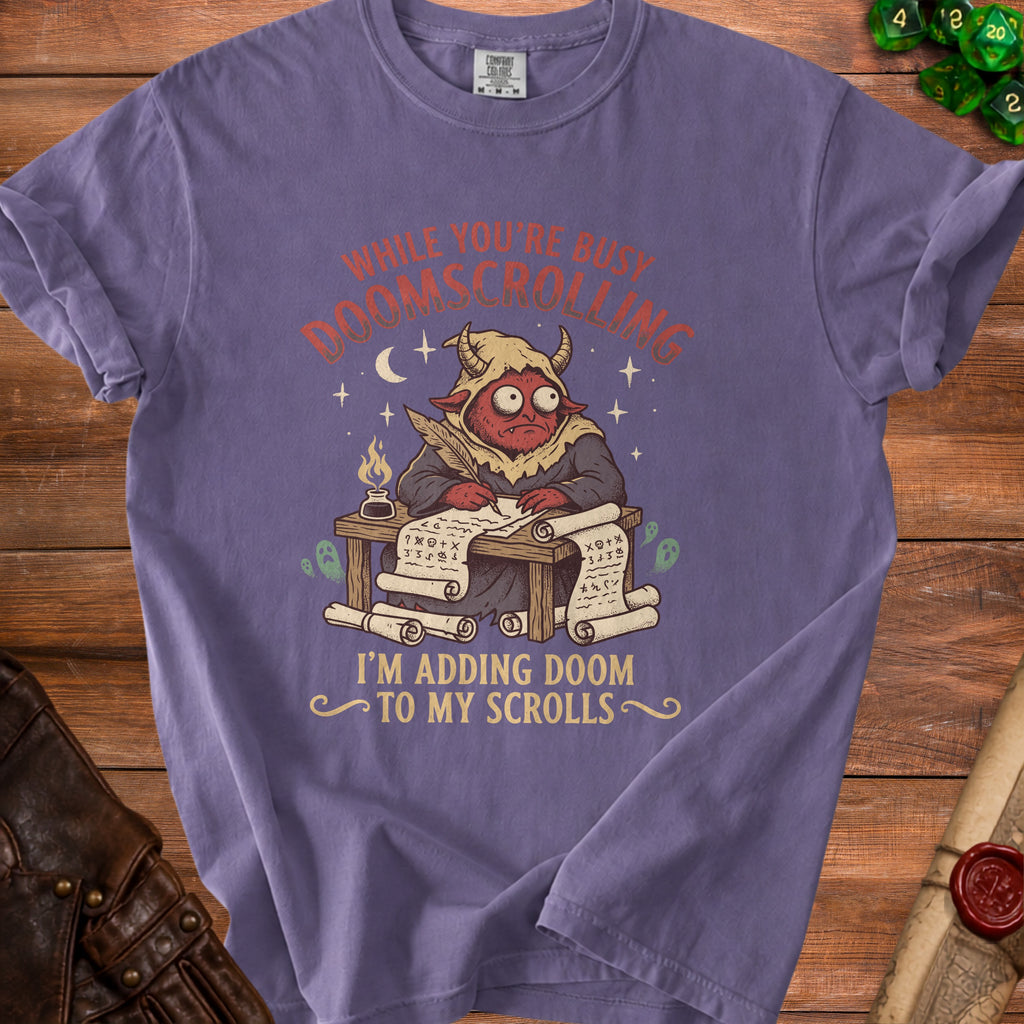 Purple t-shirt with a red demon writing on scrolls. Text reads: "While you're busy doomscrolling, I'm adding doom to my scrolls."