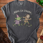T-shirt with "ABRA CA STABRA" text above a cartoon goblin and a fleeing person.