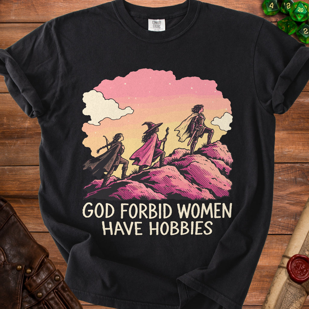 A black t-shirt features an illustration of three female adventurers climbing a pink hill, with the text GOD FORBID WOMEN HAVE HOBBIES underneath.
