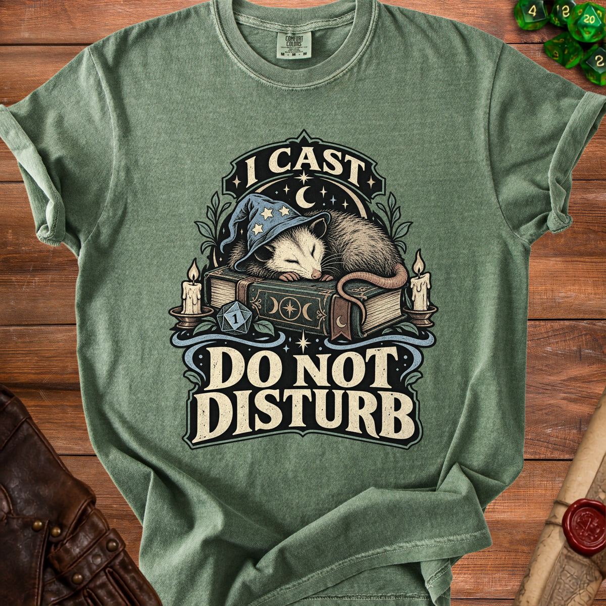 A green t-shirt with a graphic of an opossum sleeping on a book, wearing a wizard hat, with text reading "I CAST DO NOT DISTURB".