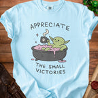 Light blue t-shirt with a cartoon goblin in a bathtub saying "Appreciate The Small Victories."