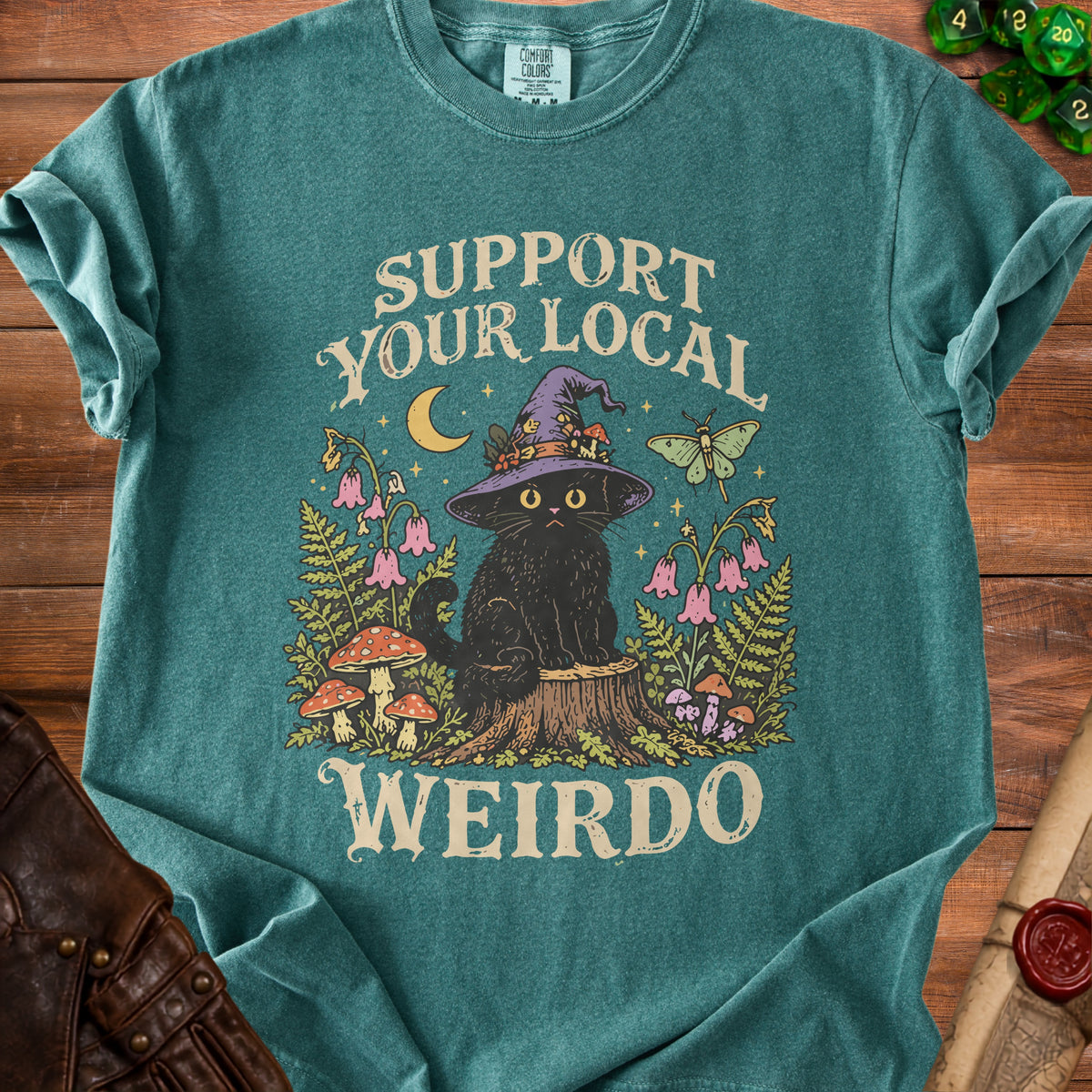 Teal crewneck t-shirt featuring a graphic of a black cat in a witch hat sitting on a tree stump, surrounded by mushrooms, flowers, and the text "SUPPORT YOUR LOCAL WEIRDO."