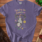 Purple t-shirt with opossum hanging from branch text "Hanging On Somehow" and "And That's Enough".