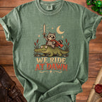 Green t-shirt with illustration of raccoon riding alligator, holding sword, text "WE RIDE AT DAWN".