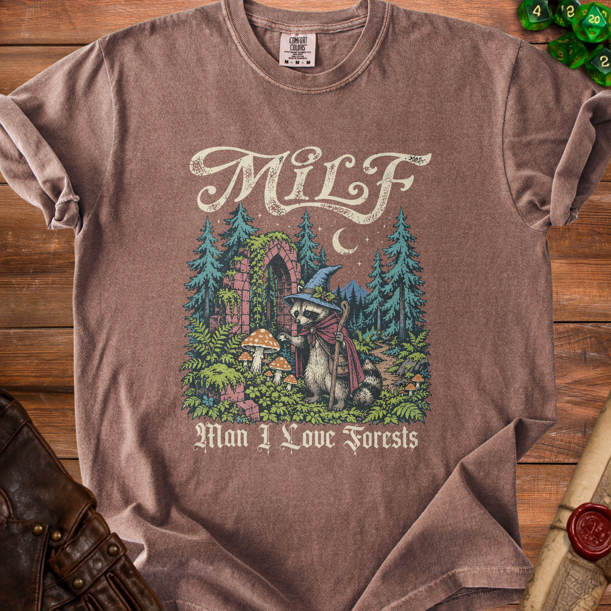 Brown t-shirt with a raccoon in a witch hat and cape, surrounded by trees and mushrooms.