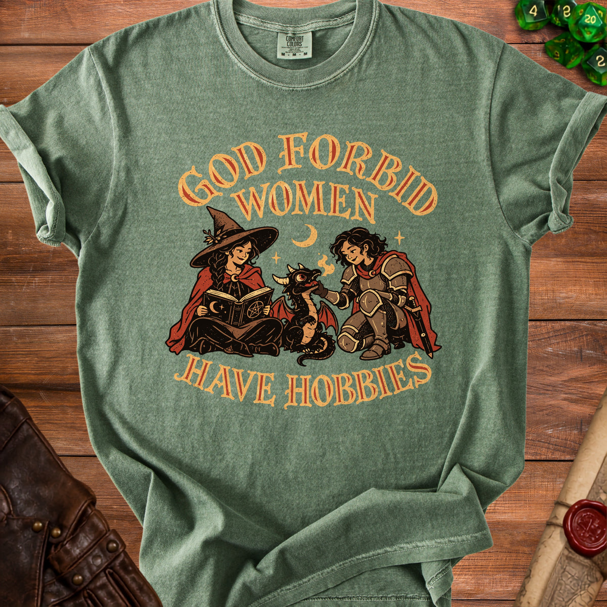 Sage green t-shirt featuring a retro graphic of a witch, knight, and dragon with text reading "God Forbid Women Have Hobbies".