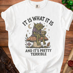 White t-shirt graphic: goblin on mushroom, holding coffee, with text "IT IS WHAT IT IS AND IT'S PRETTY TERRIBLE".