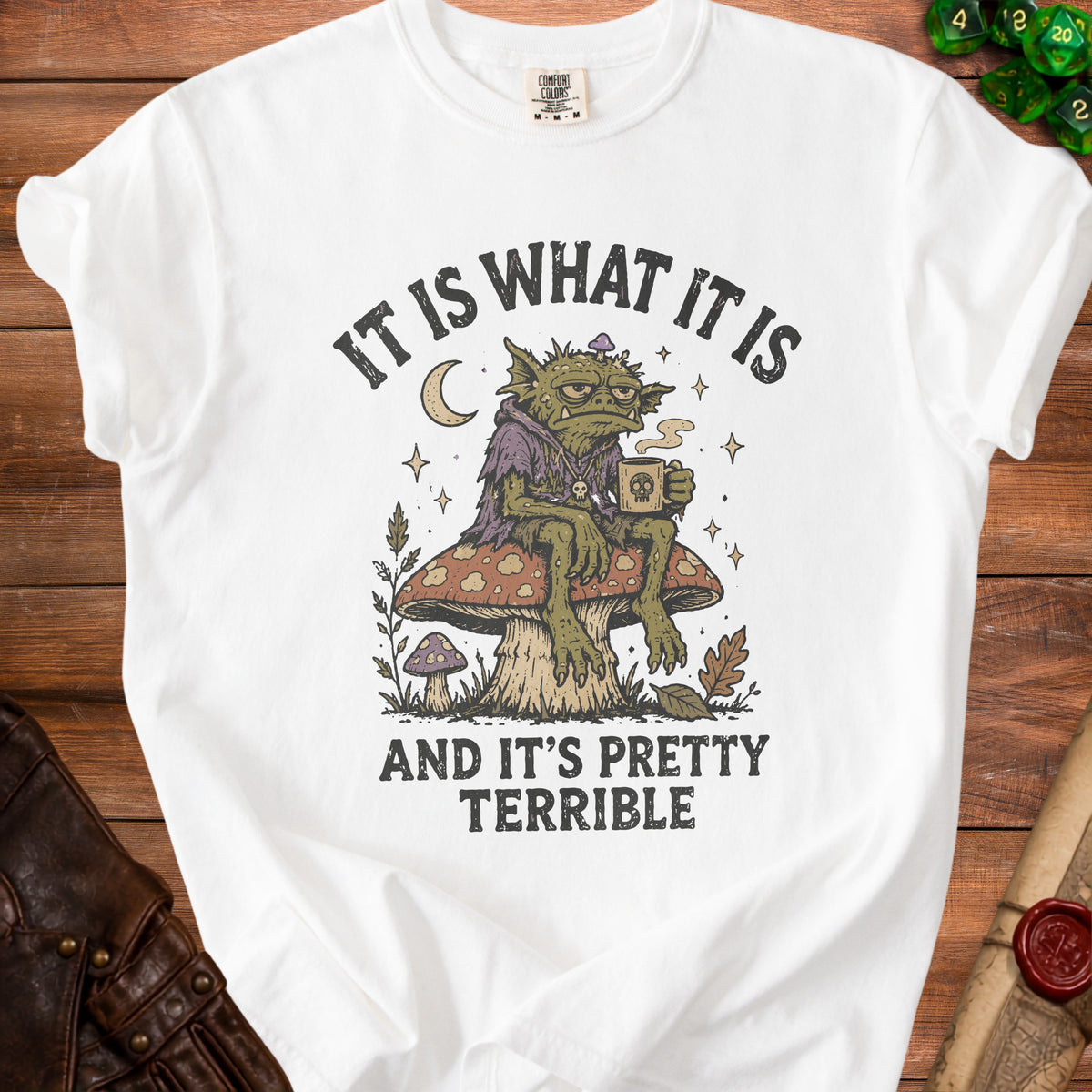 White t-shirt graphic: goblin on mushroom, holding coffee, with text "IT IS WHAT IT IS AND IT'S PRETTY TERRIBLE".