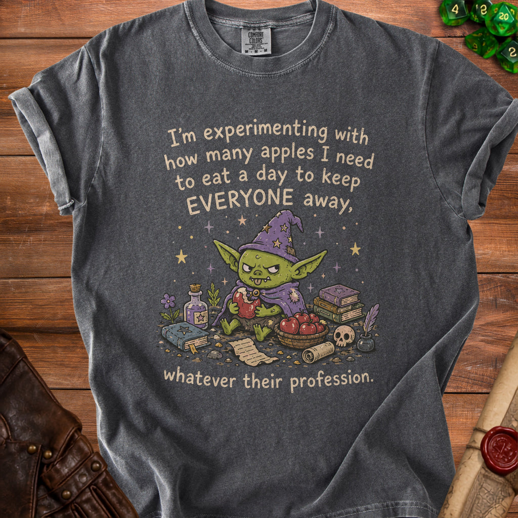 Dark grey t-shirt with a goblin wearing a witch hat holding an apple. Text reads "I'm experimenting with how many apples I need to eat a day to keep EVERYONE away, whatever their profession."