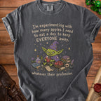 Dark grey t-shirt with a goblin wearing a witch hat holding an apple. Text reads "I'm experimenting with how many apples I need to eat a day to keep EVERYONE away, whatever their profession."