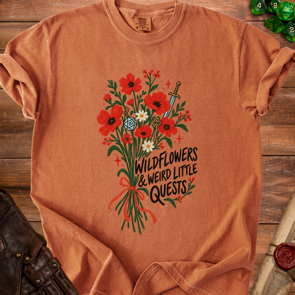 T-shirt with wildflowers, sword, key, d20, and text "Wildflowers & Weird Little Quests".