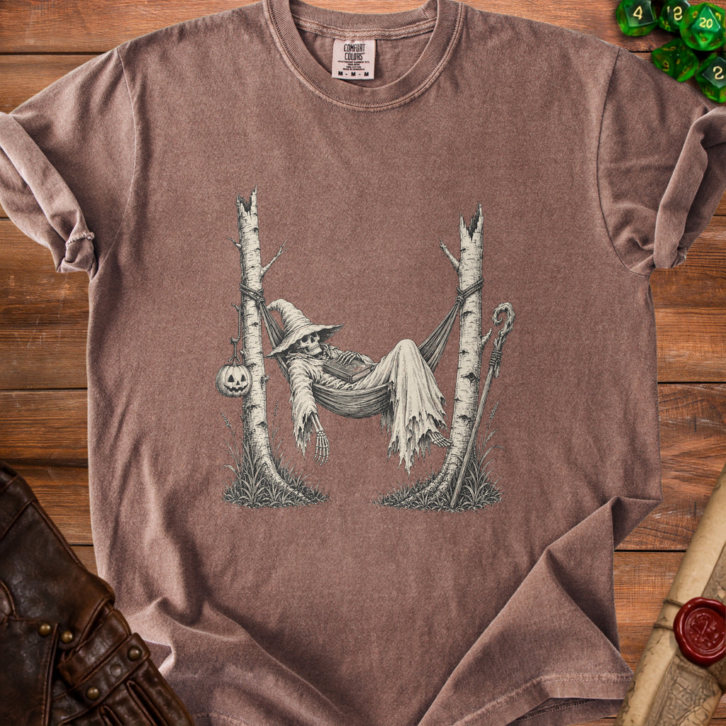 T-shirt graphic of skeleton in witch hat resting in hammock between trees with pumpkin.