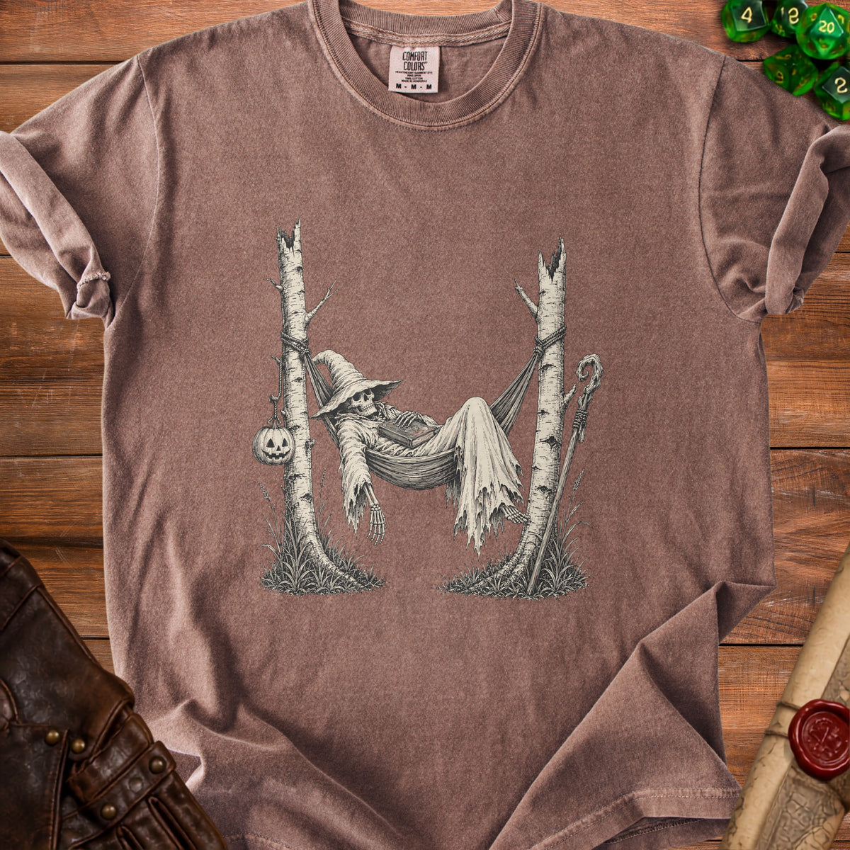 T-shirt graphic of skeleton in witch hat resting in hammock between trees with pumpkin.