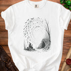 White t-shirt with a black graphic of a witch drinking from a mug near a cave entrance with bats flying.