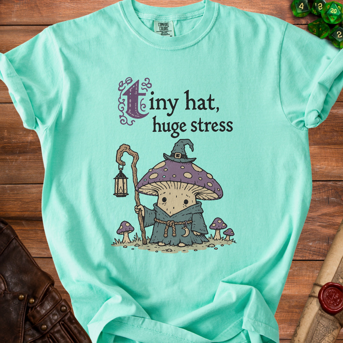 Mint green t-shirt with illustration of a mushroom character and text "tiny hat, huge stress".