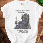 White t-shirt with skeleton in wizard robe, mug, books, and text "SOCIAL BATTERY DRAINED PLEASE CAST LEFT ALONE".