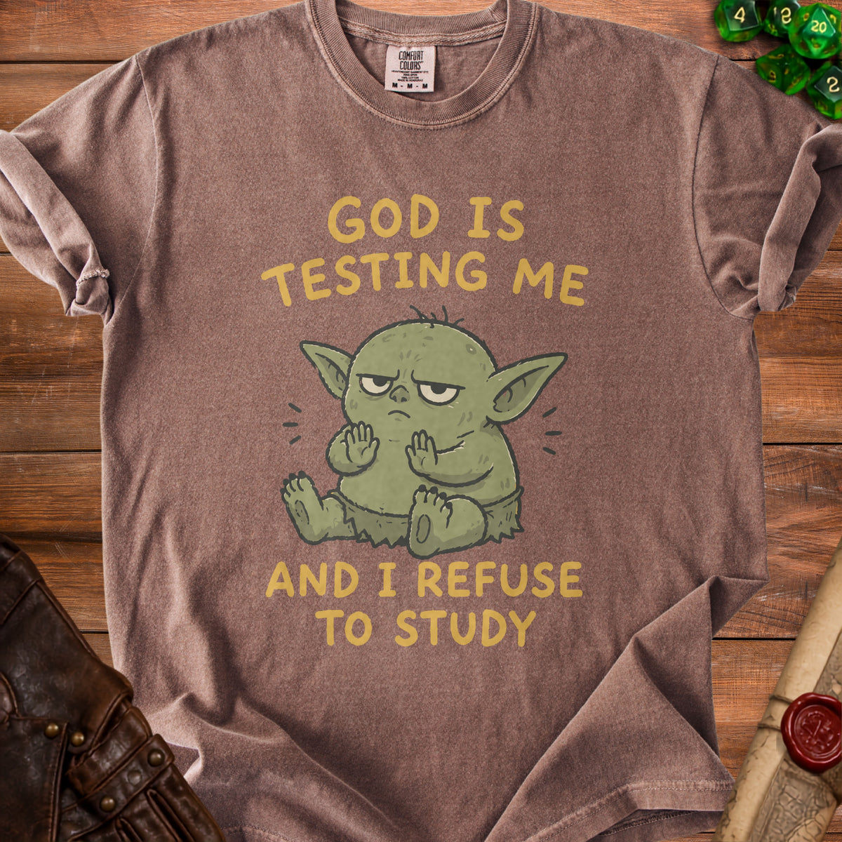 Brown t-shirt with a green cartoon character and yellow text reading "GOD IS TESTING ME AND I REFUSE TO STUDY".