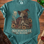Teal t-shirt featuring a skeletal necromancer surrounded by flowers, holding a book, and text reading Necromancer in Bloom.