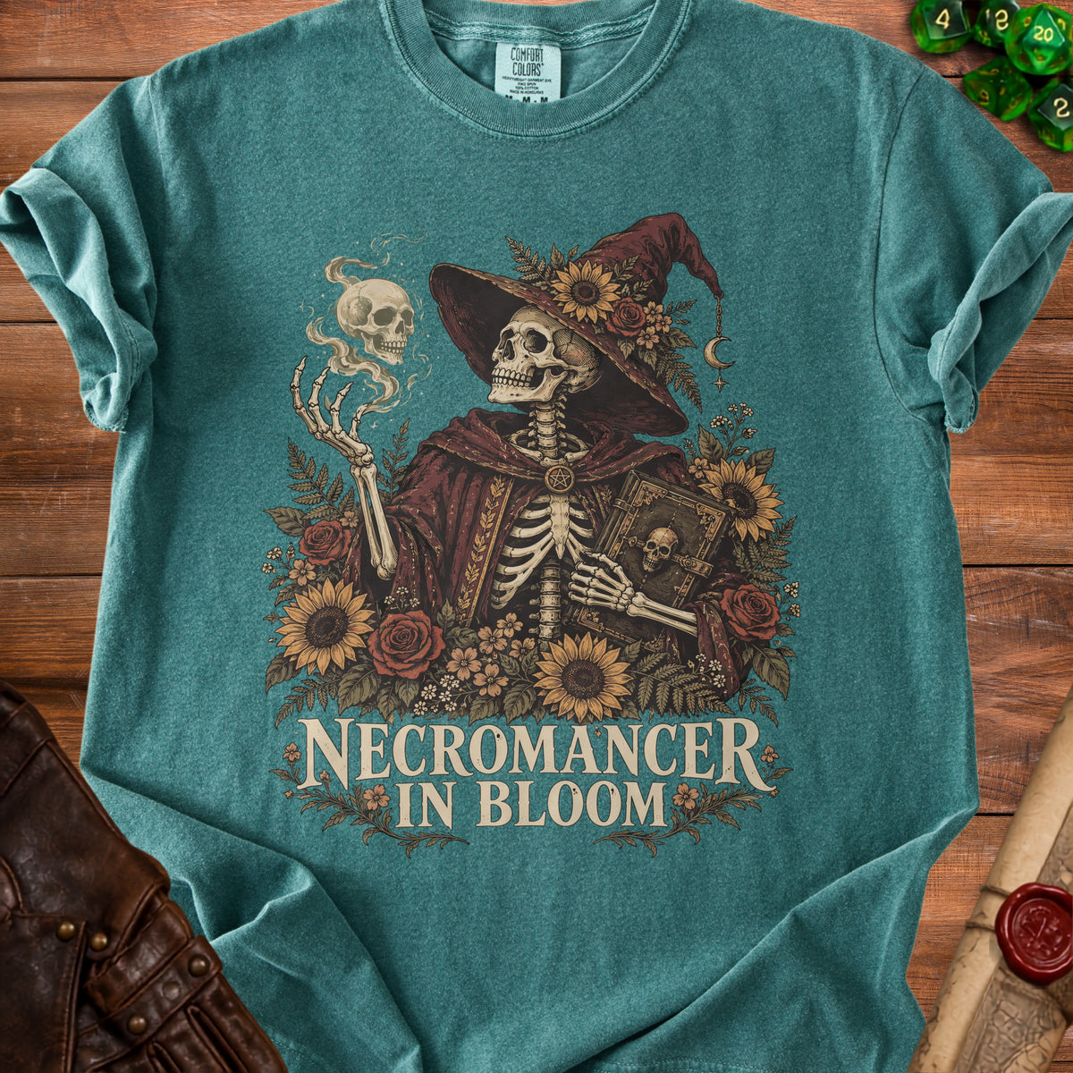 Teal t-shirt featuring a skeletal necromancer surrounded by flowers, holding a book, and text reading Necromancer in Bloom.