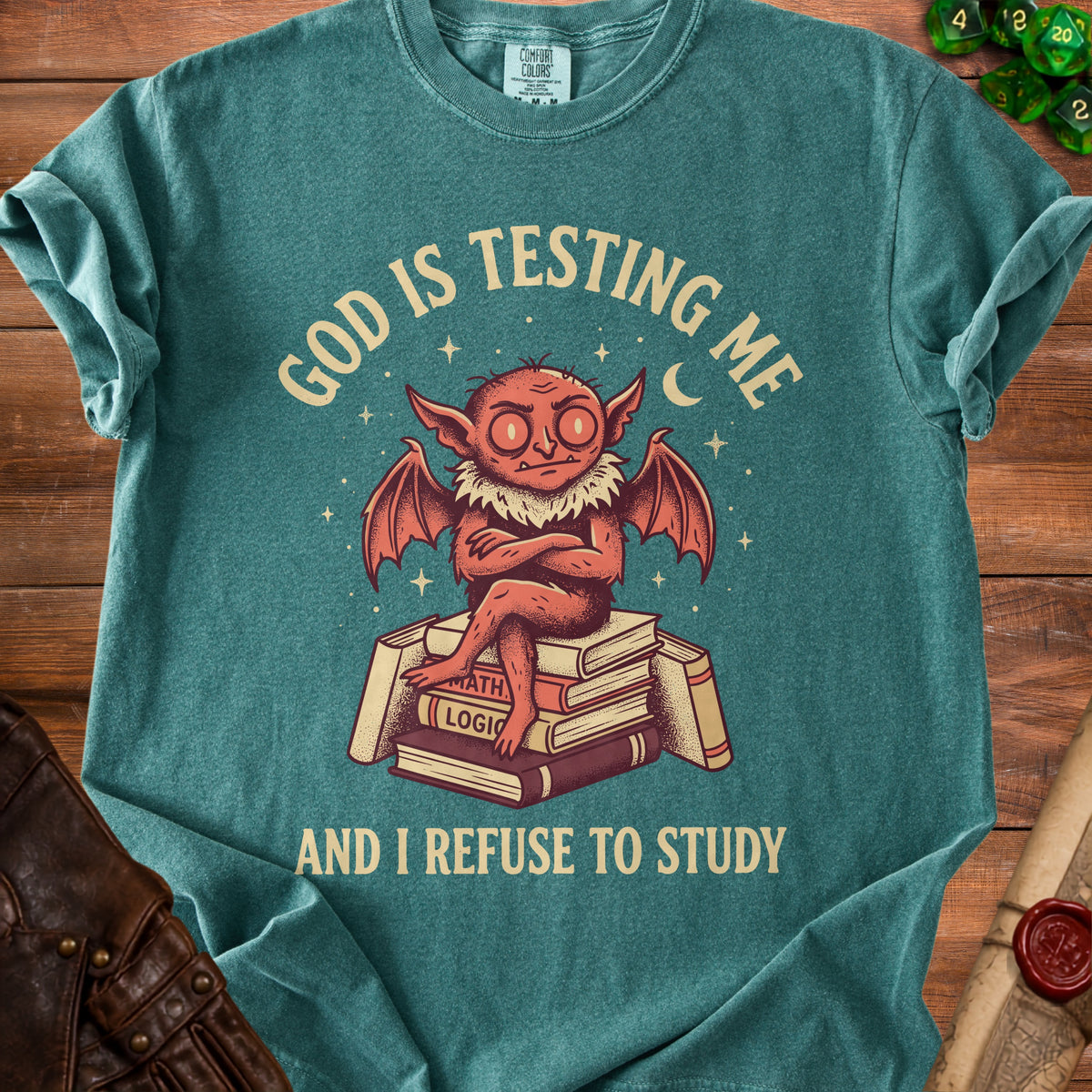 Teal t-shirt with a red creature sitting on books, text reads "God is testing me and I refuse to study".