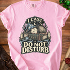 A light pink t-shirt displays graphic text reading I Cast Do Not Disturb above a sleeping possum wearing a wizard hat, resting on a spellbook flanked by candles.