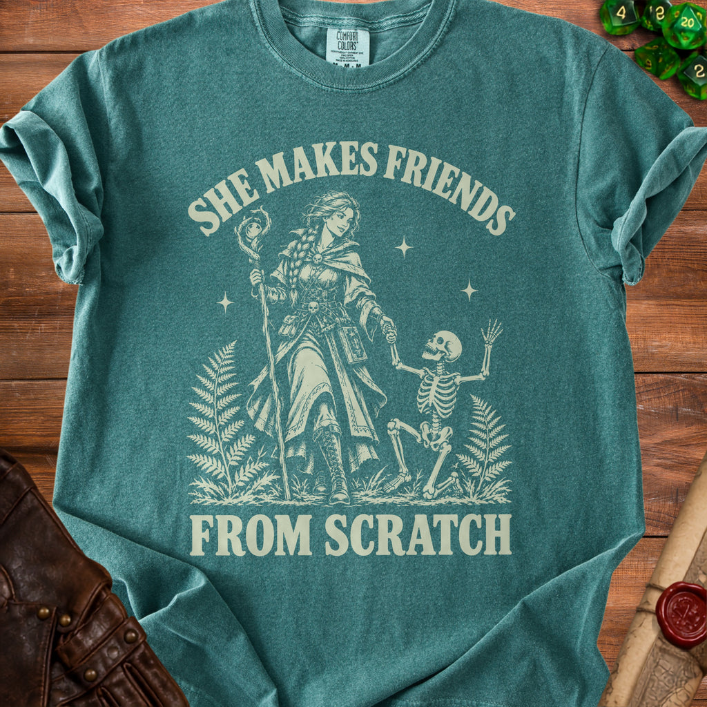 Teal t-shirt with a graphic of a woman with a staff and a skeleton, and text "SHE MAKES FRIENDS FROM SCRATCH".