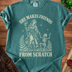 Teal t-shirt with a graphic of a woman with a staff and a skeleton, and text "SHE MAKES FRIENDS FROM SCRATCH".