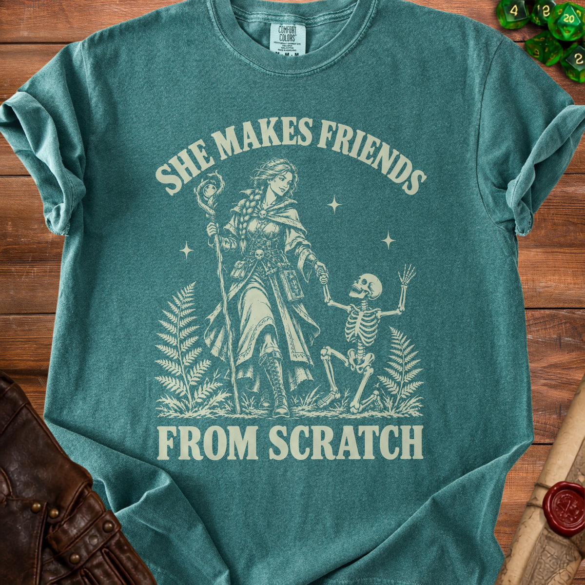 Teal t-shirt with a graphic of a woman with a staff and a skeleton, and text "SHE MAKES FRIENDS FROM SCRATCH".