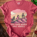 Dusty rose t-shirt featuring three female adventurers climbing a hill, with the text "God forbid women have hobbies."