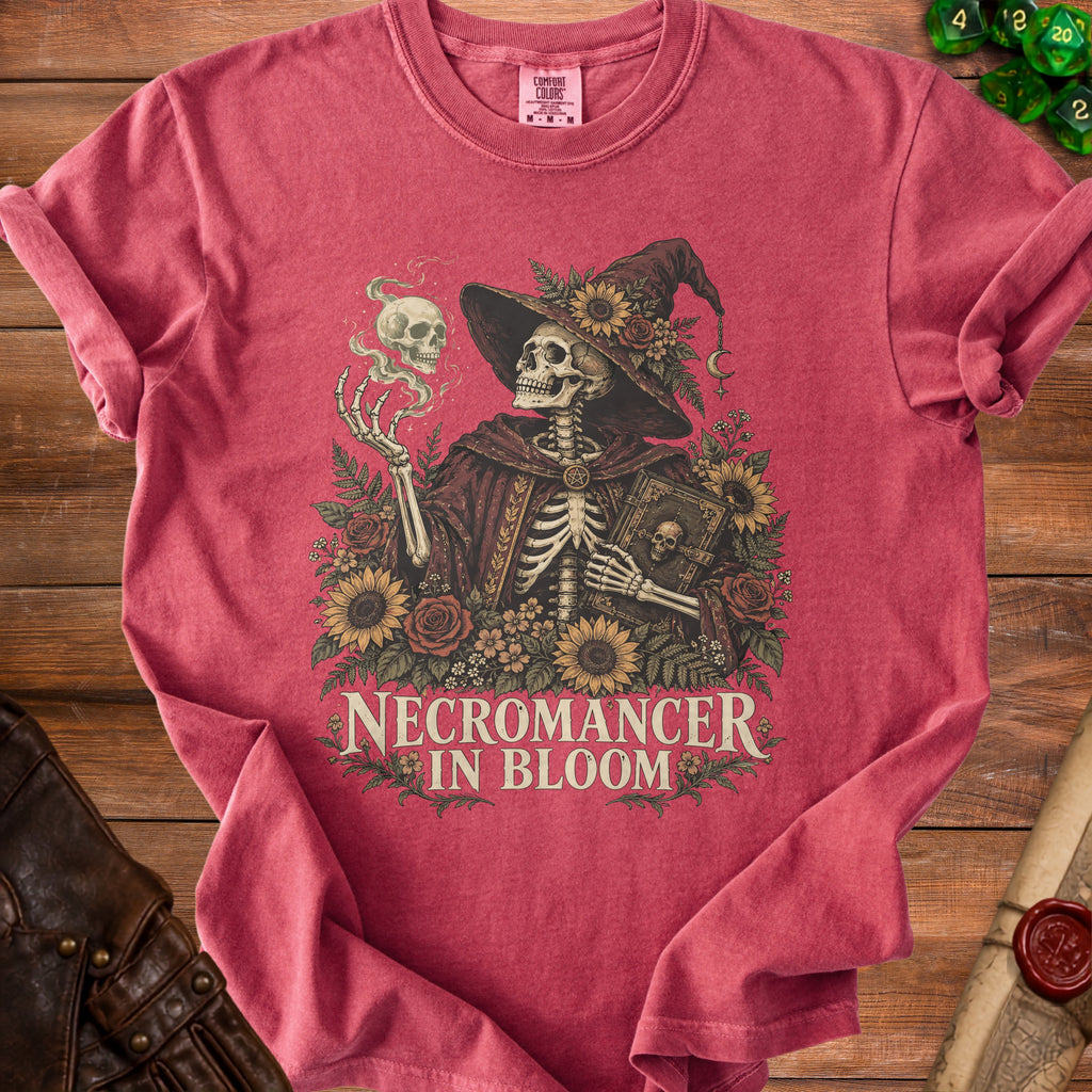 Red t-shirt featuring a skeleton in a witch hat and robe, holding a skull and book, with floral elements and the text "NECROMANCER IN BLOOM."