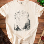 T-shirt with a black graphic of a witch drinking tea next to a cave and swirling bats.