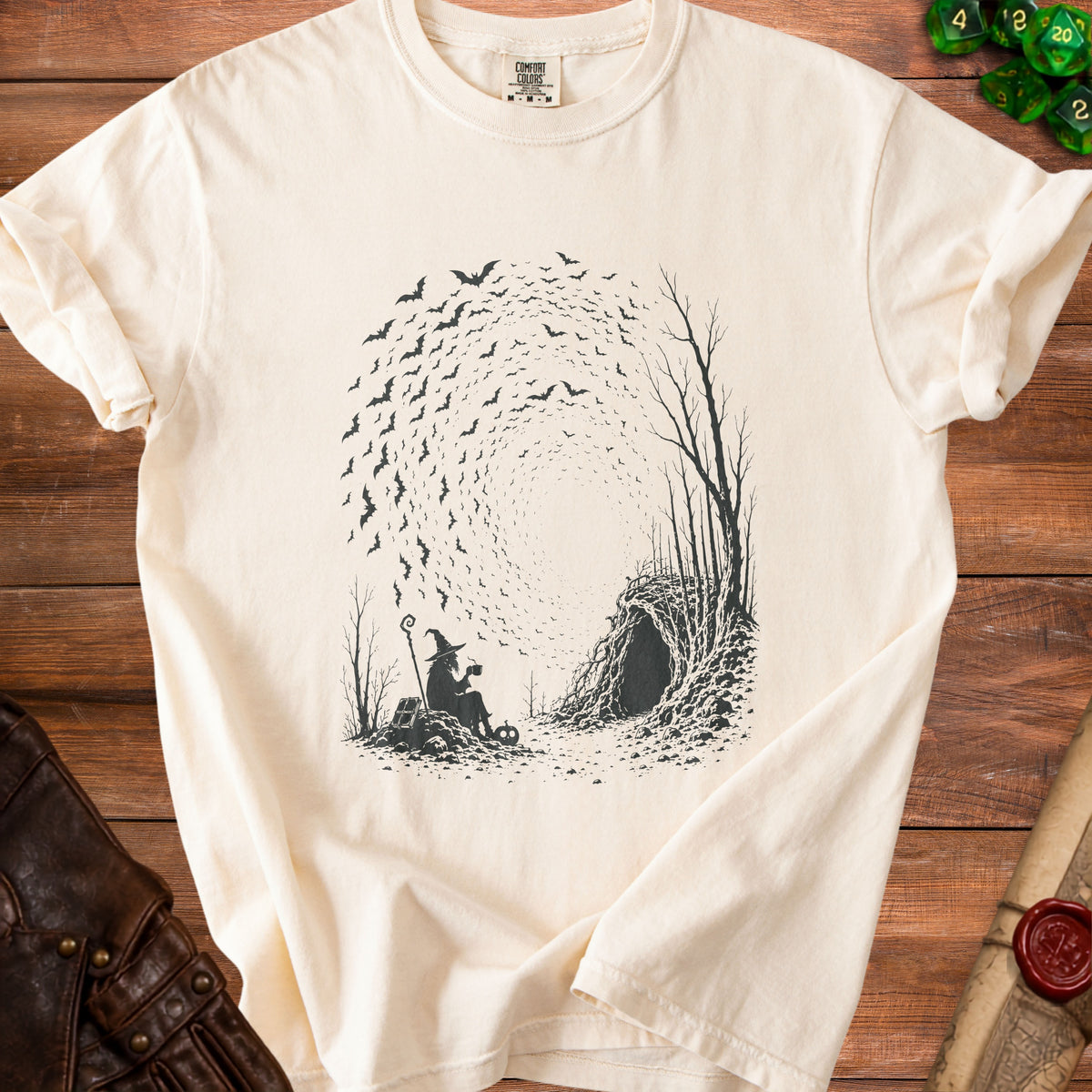 T-shirt with a black graphic of a witch drinking tea next to a cave and swirling bats.