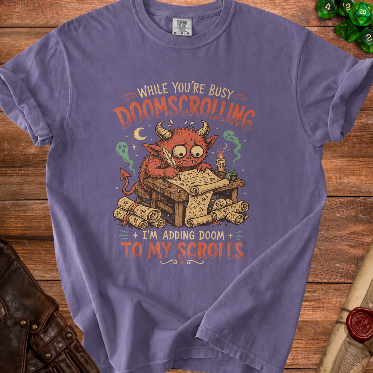Purple t-shirt with a cartoon devil adding doom to scrolls with the text "WHILE YOU'RE BUSY DOOMSCROLLING + I'M ADDING DOOM + TO MY SCROLLS".