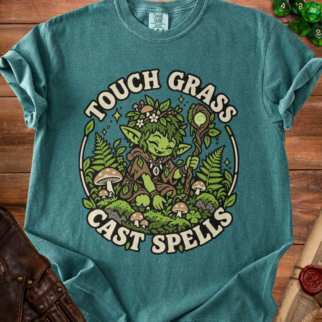 Green t-shirt with illustration of a gnome surrounded by plants, mushrooms, and text "Touch Grass Cast Spells."