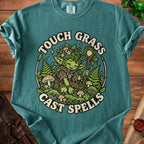 Green t-shirt with illustration of a gnome surrounded by plants, mushrooms, and text "Touch Grass Cast Spells."