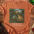 Orange t-shirt with three frogs in forest and text "Strange But Excellent Company".