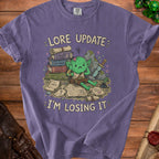 Purple t-shirt with a cartoon goblin overwhelmed by books and scrolls with "Lore Update" and "I'm Losing It".