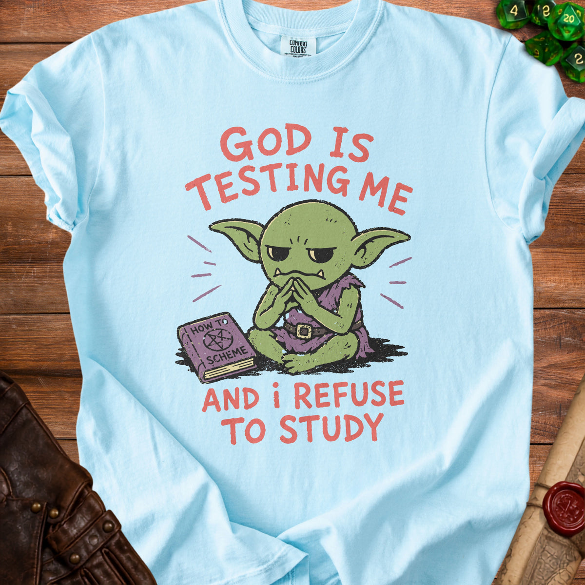 Light blue t-shirt with green goblin sitting by a book titled "How to Scheme." Text reads "God is testing me and I refuse to study."