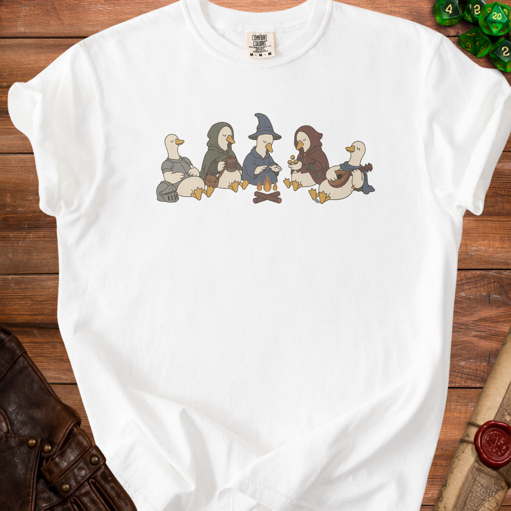 White t-shirt with an illustration of five anthropomorphic geese in fantasy costumes around a campfire.