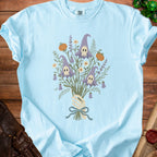Light blue t-shirt with a skeleton hand holding a bouquet of witch hats, skulls, pumpkins, and flowers.