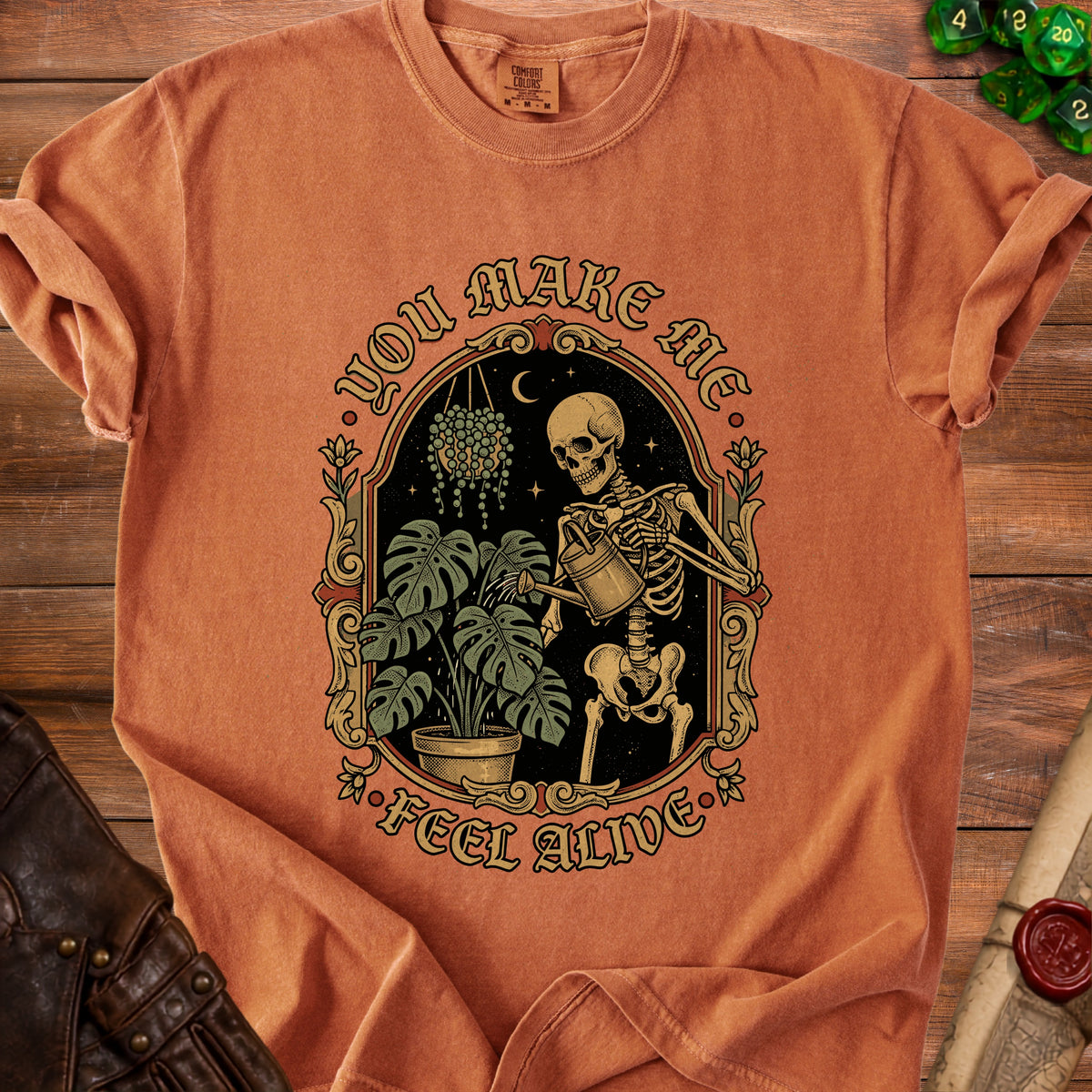Orange t-shirt with skeleton watering plants and "You Make Me Feel Alive" text.