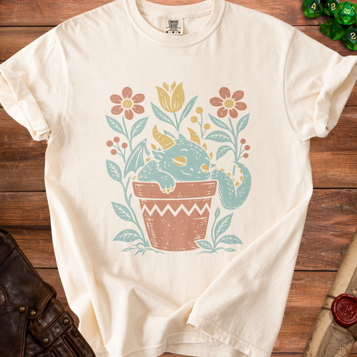 Cream t-shirt with a faded teal and coral dragon sleeping in a terracotta pot with flowers.