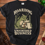 Black t-shirt with a distressed graphic of a dragon resting on scrolls with text "Hoarding Unfinished Business".
