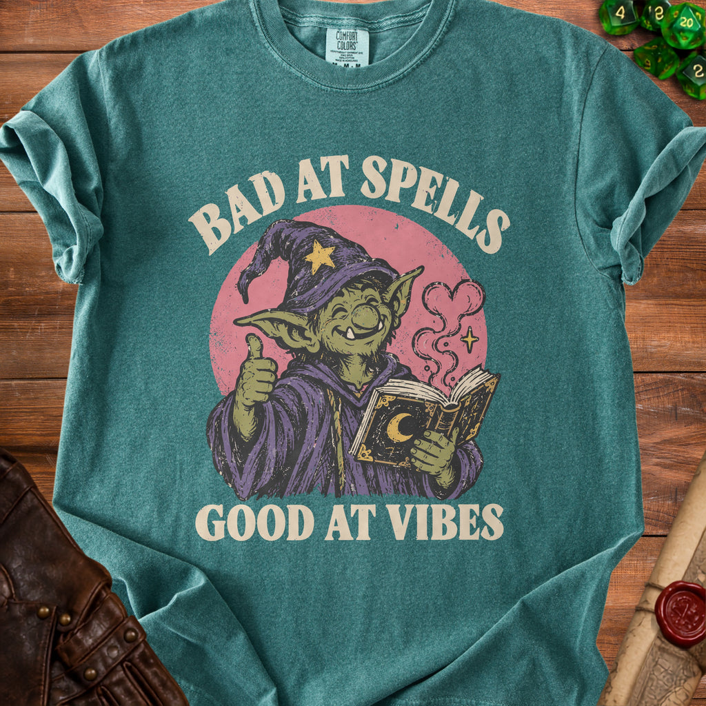 Green t-shirt with graphic of a goblin wizard holding a book, "Bad at Spells Good at Vibes".