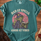 Green t-shirt with graphic of a goblin wizard holding a book, "Bad at Spells Good at Vibes".