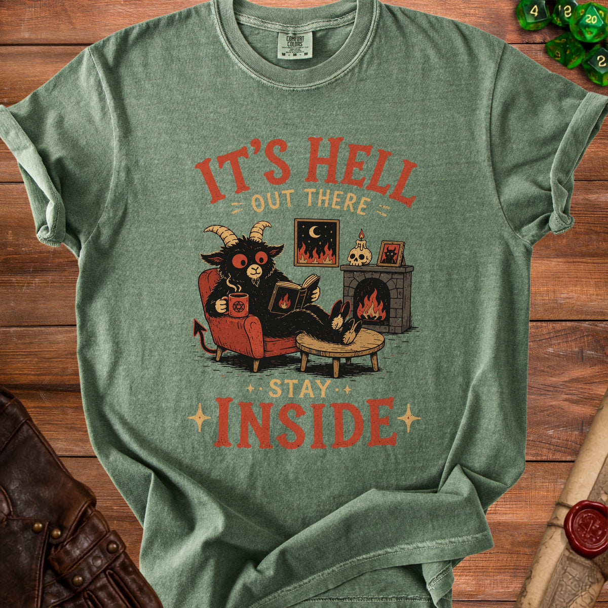 Green t-shirt with "It's Hell Out There Stay Inside" text and a devil reading by a fireplace.
