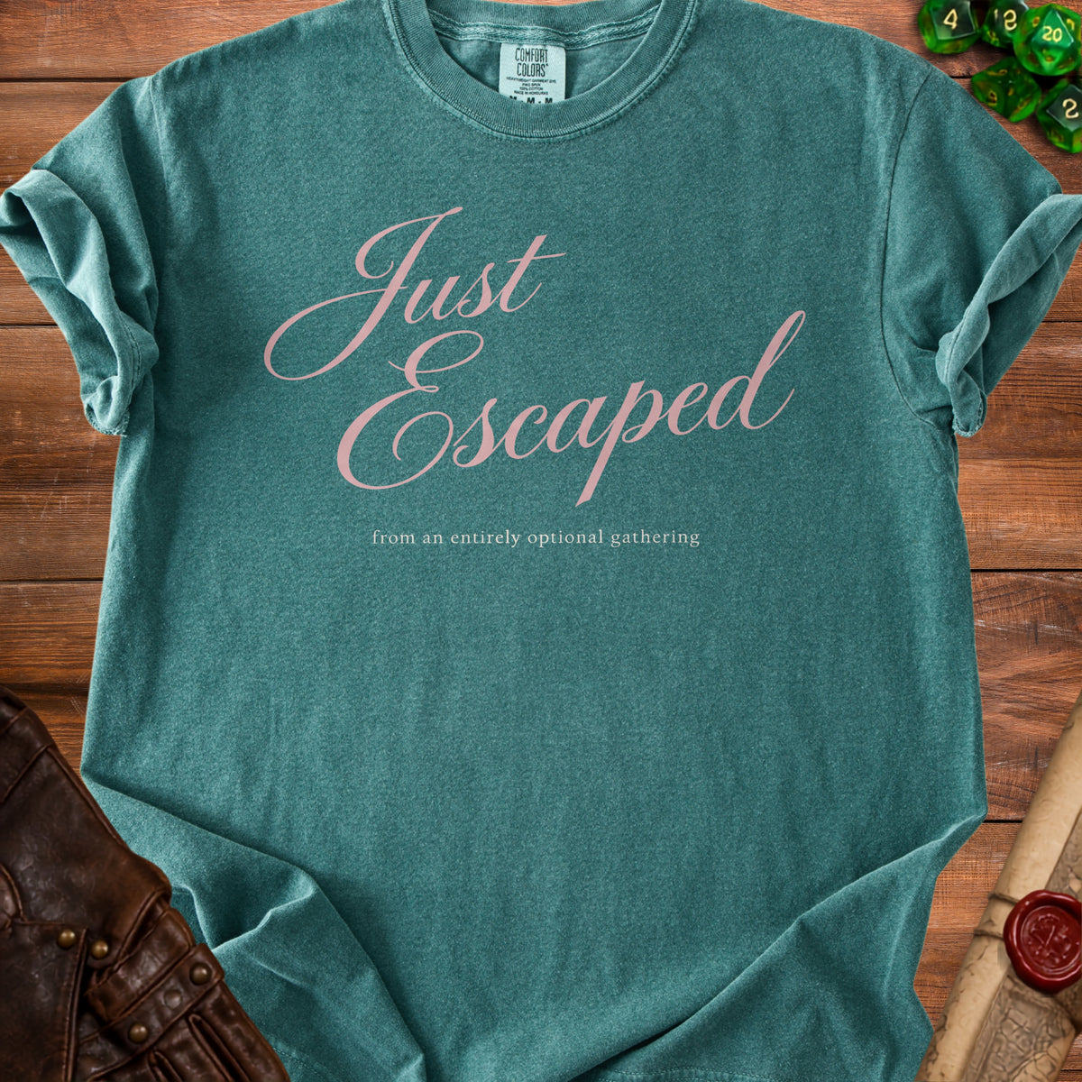 A teal t-shirt with pink script text reading Just Escaped from an entirely optional gathering on a wooden background.