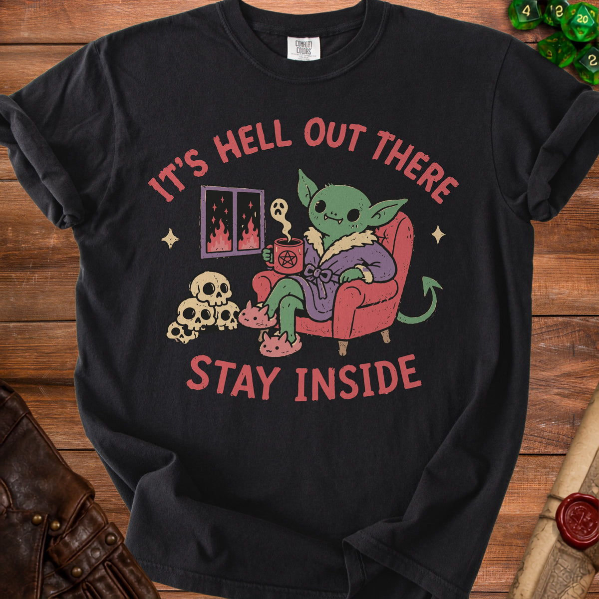 Black t-shirt with a graphic of a goblin sitting in a chair with text "It's Hell Out There Stay Inside".