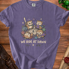 Purple t-shirt featuring a graphic of cartoon owls playing a tabletop role-playing game with dice and text "WE RIDE AT DAWN".