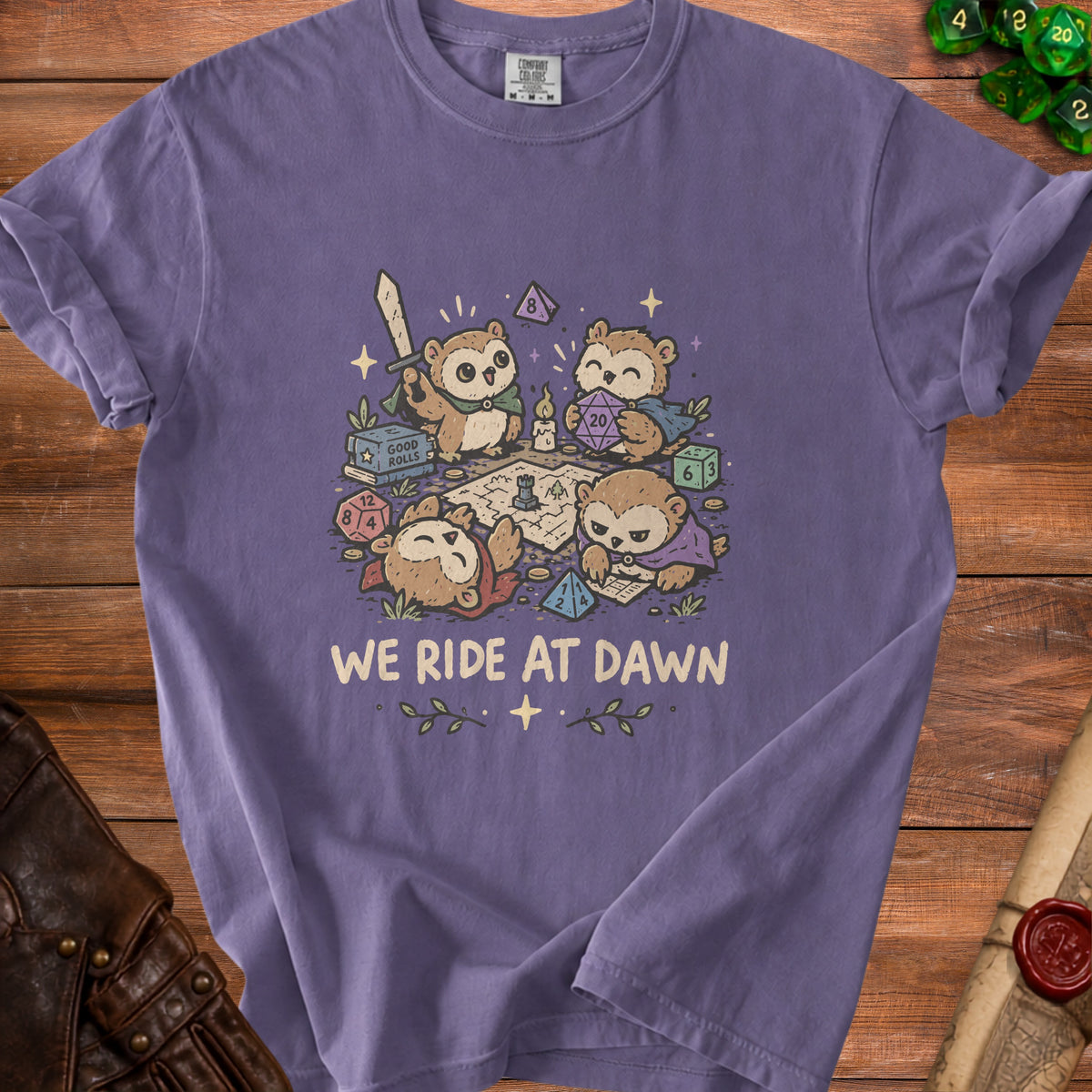 Purple t-shirt featuring a graphic of cartoon owls playing a tabletop role-playing game with dice and text "WE RIDE AT DAWN".